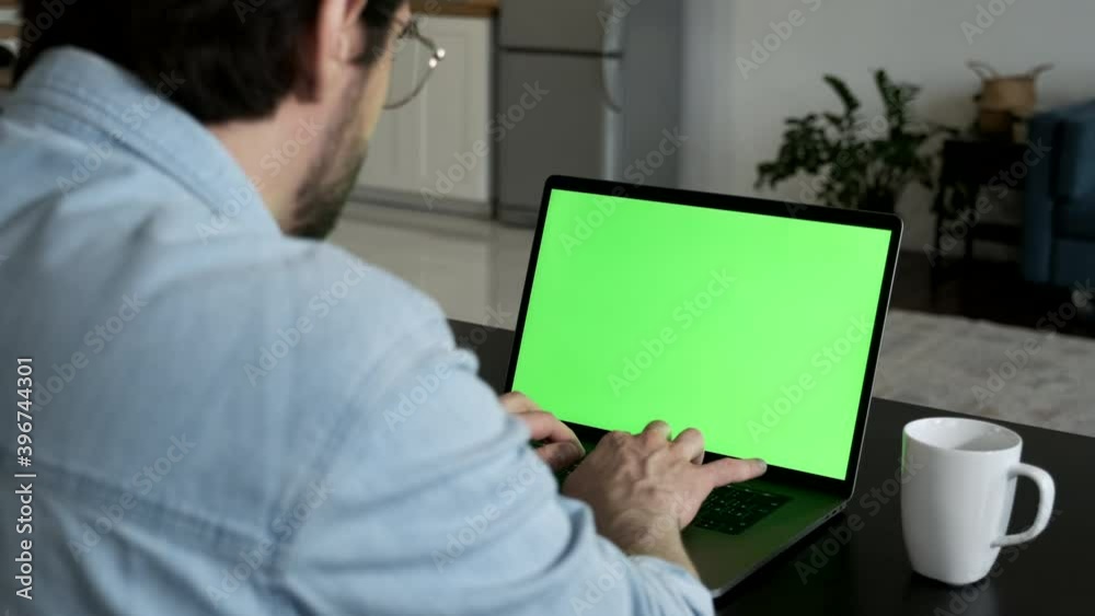 Over the Shoulder Camera Shot of Man at Home Sitting on a Couch Working on Green Mock-up Screen Laptop Computer Online 