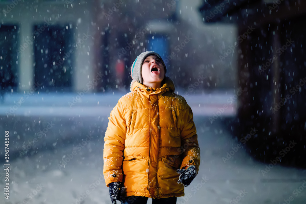 Little school kid boy of elementary class walking to school during snowfall on early dark morning. Happy healthy child having fun and playing with snow. Schoolkid in colorful winter clothes.