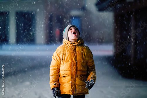 Little school kid boy of elementary class walking to school during snowfall on early dark morning. Happy healthy child having fun and playing with snow. Schoolkid in colorful winter clothes.