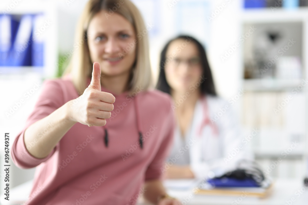 Patient holds thumb up in medical office in background doctor sits. Patient holds thumb up in medical office in background doctor sits.