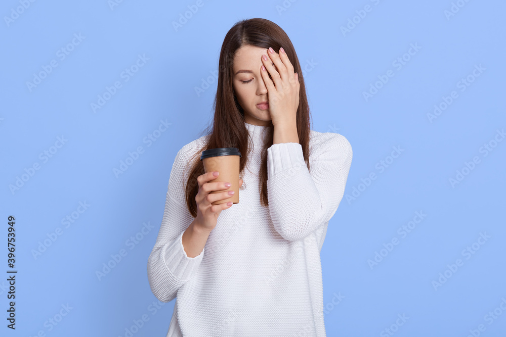 Displeased sleepy tired woman covers face with one palm, drinks fresh coffee or cappuccino, wears white jumper, has unhappy look, poses over blue background. Displeased sleepy tired woman covers face with one palm, drinks fresh coffee or cappuccino, wears white jumper, has unhappy look, poses over blue background.