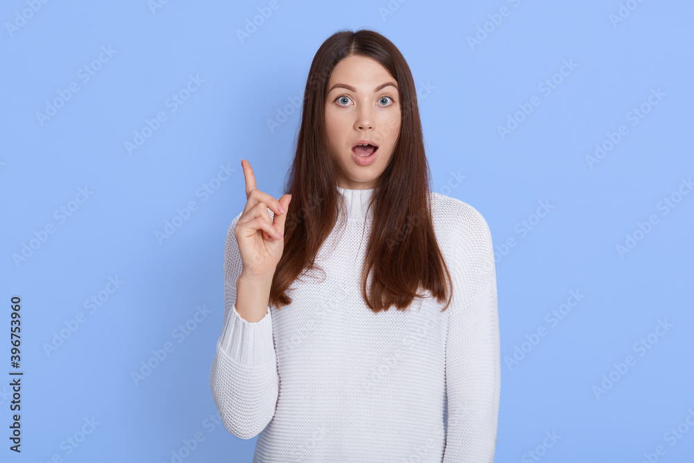 Amazed shocked young woman pointing up with fore finger, keeps mouth widely opened, wearing casual white shirt, has dark hair, being shocked and astonished, stands against blue wall. Amazed shocked young woman pointing up with fore finger, keeps mouth widely opened, wearing casual white shirt, has dark hair, being shocked and astonished, stands against blue wall.