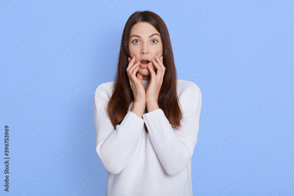 Stunned caucasian female stares at camera with big eyes and keeps mouth opened, looks surprisingly, realizes bad news, stands against blue background, keeps fingers on cheeks. Stunned caucasian female stares at camera with big eyes and keeps mouth opened, looks surprisingly, realizes bad news, stands against blue background, keeps fingers on cheeks.
