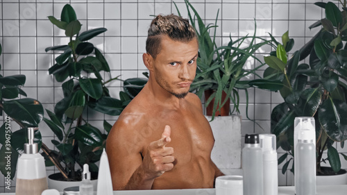 young shirtless man pointing with finger while looking at camera near green plants on blurred background in bathroom