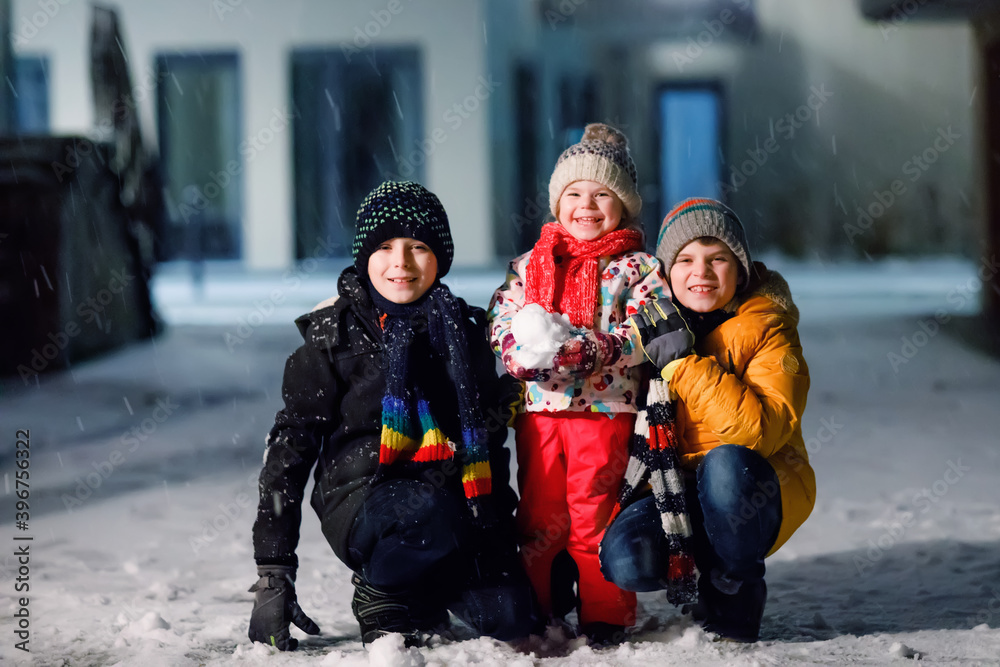Two little kid boys and cute toddler girl sitting together playing with snow on winter night. Siblings, brothers and baby sister enjoying strong snowfall. Active fun for family vacation