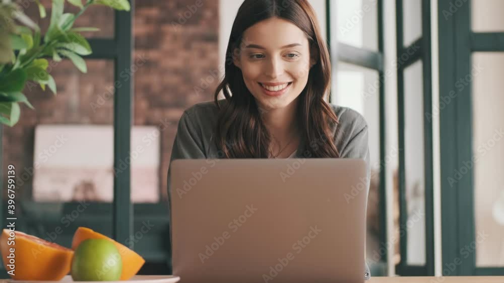 Positive happy young woman sitting at the kitchen and using laptop computer indoors at home