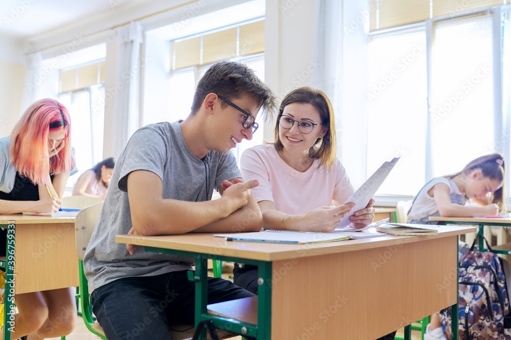 Lesson in class of high school students, female teacher sitting at desk with student Lesson in class of high school students, female teacher sitting at desk with student