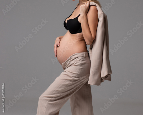 Young beautiful woman with blond hair is pregnant in a white fashionable elegant trouser suit sits on a chair on a gray background. Fashion for pregnant women. Business woman is pregnant. 