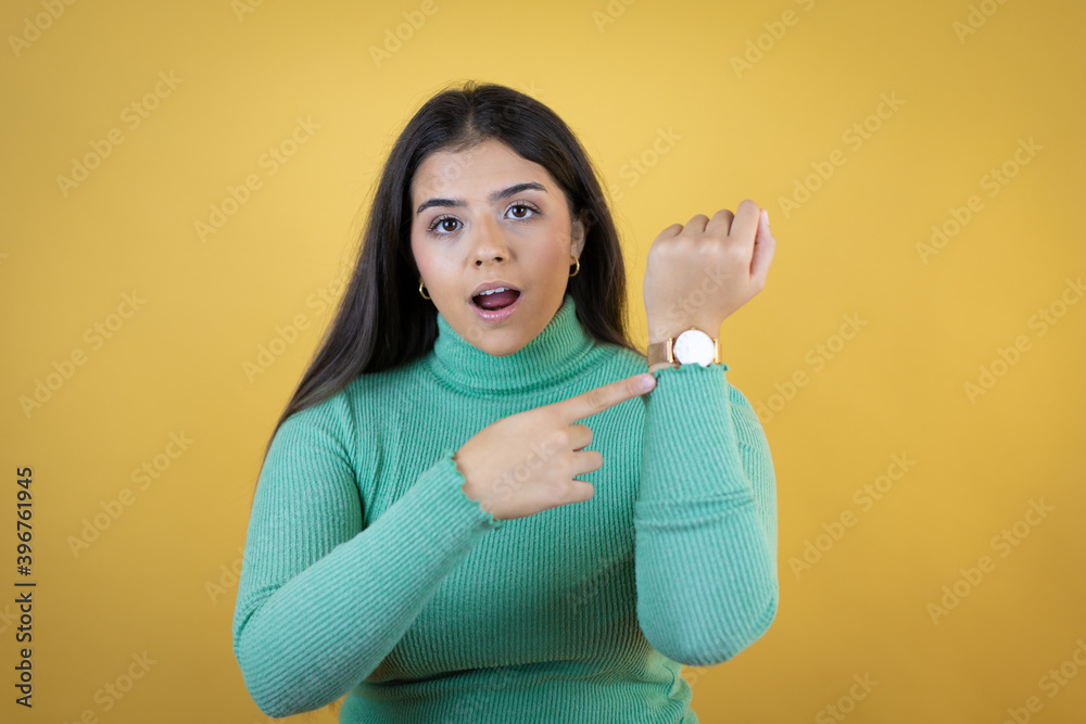 Obraz premium Young caucasian woman over isolated yellow background surprised and pointing her watch because it's late