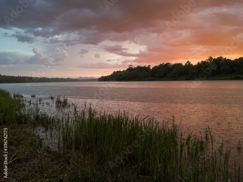 Landscape with river  colorful clouds  tall water grass and mountain in distance at sunset. Dramatic riverine scene during cloudy dusk with rain or summer shower and majestic beautiful glow in clouds