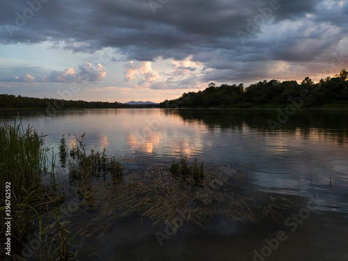 Landscape with river, dark clouds, tall water grass and mountain in distance at sunset. Calm riverine scene during cloudy evening.