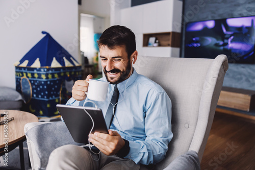 Young bearded father sitting in chair i living room, drinking his morning coffee and using tablet during quarantine. Covid 19 outbreak concept. photo