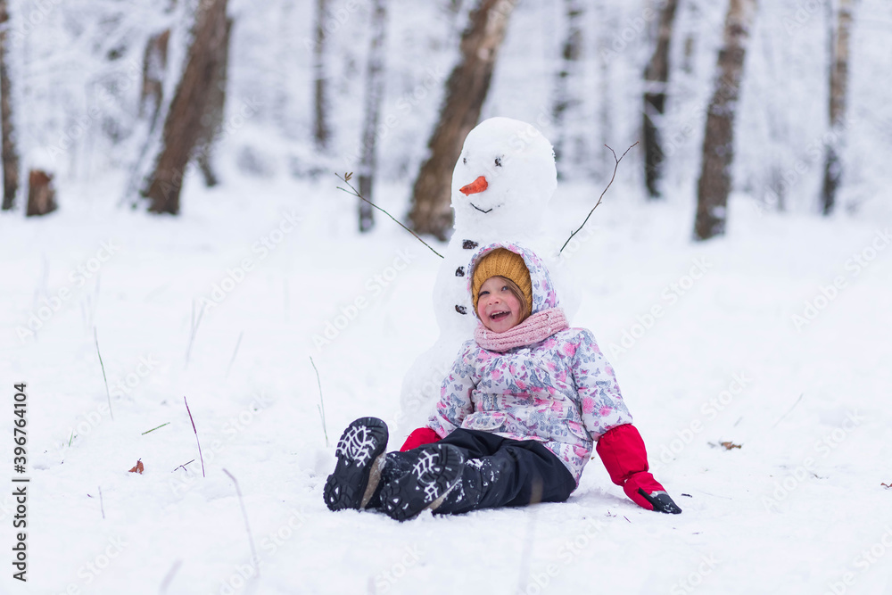 
Happy child plays with a snowman on a winter walk in nature in the forest. Funny little girl laughs and sits on the snow next to a snowman in winter outdoors
