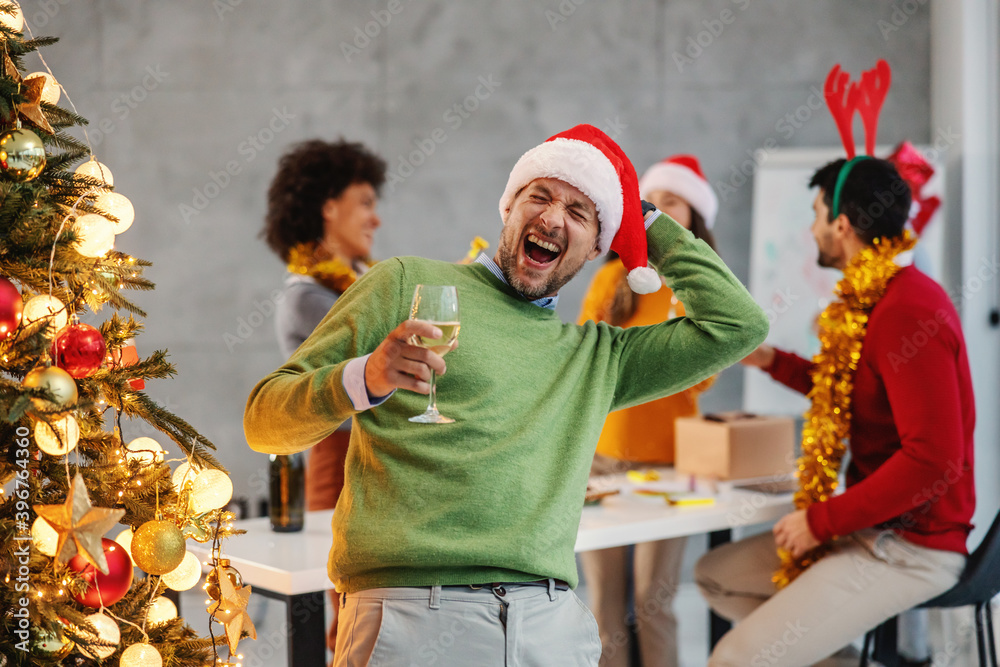 Young overjoyed businessman with santa's hat holding champagne while standing next to christmas tree in his firm on christmas eve. In background are his colleagues celebrating, too.