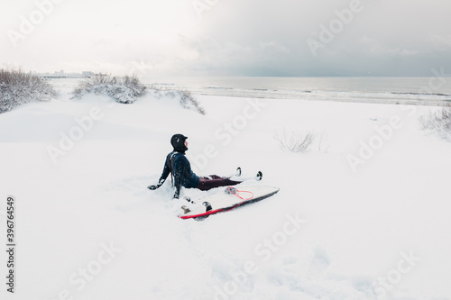 Cold winter and surfer sitting on snow beach with surfboard. Winter with surfer in wetsuit.