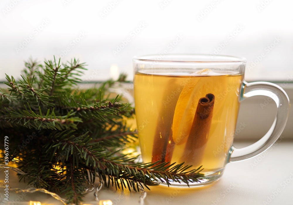 Hot winter flavored drink. Glass mug with tea, orange and cinnamon on the window. Green branches of the Christmas tree Hot winter flavored drink. Glass mug with tea, orange and cinnamon on the window. Green branches of the Christmas tree