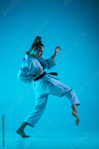 Active training. Professional female judoist in white judo kimono practicing and training isolated on blue neoned studio background. Grace of motion and action. Healthy lifestyle, sport concept