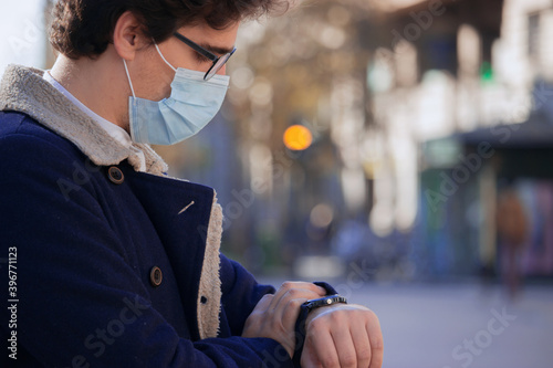 A recognizable young man wearing a mask looks at his watch on a winter day in the city center. Man is in a hurry because he is late for work.