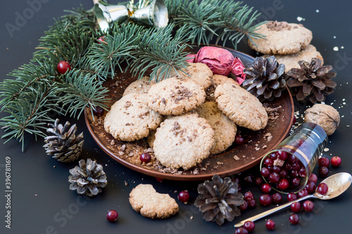 Preparing for Christmas. A plate with homemade oatmeal cookies, a fresh cranberries, a spruce branch and pine cones on a dark background. Selective focus.