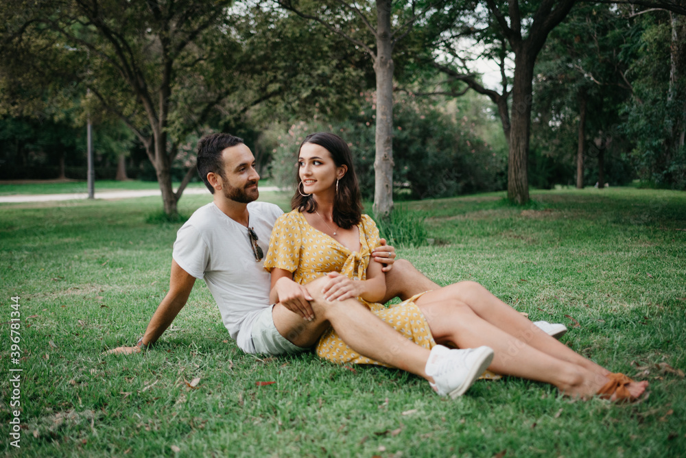 A brunette girl in a yellow dress is sitting between the legs of her boyfriend on the grass in old Spain town. A laughing couple of tourists on a date in the Valencian park. A brunette girl in a yellow dress is sitting between the legs of her boyfriend on the grass in old Spain town. A laughing couple of tourists on a date in the Valencian park.