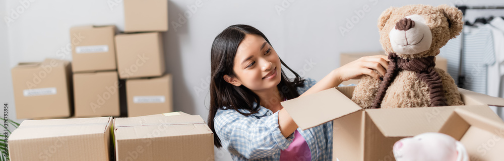 Positive asian volunteer putting soft toy in box near packages on blurred foreground, banner