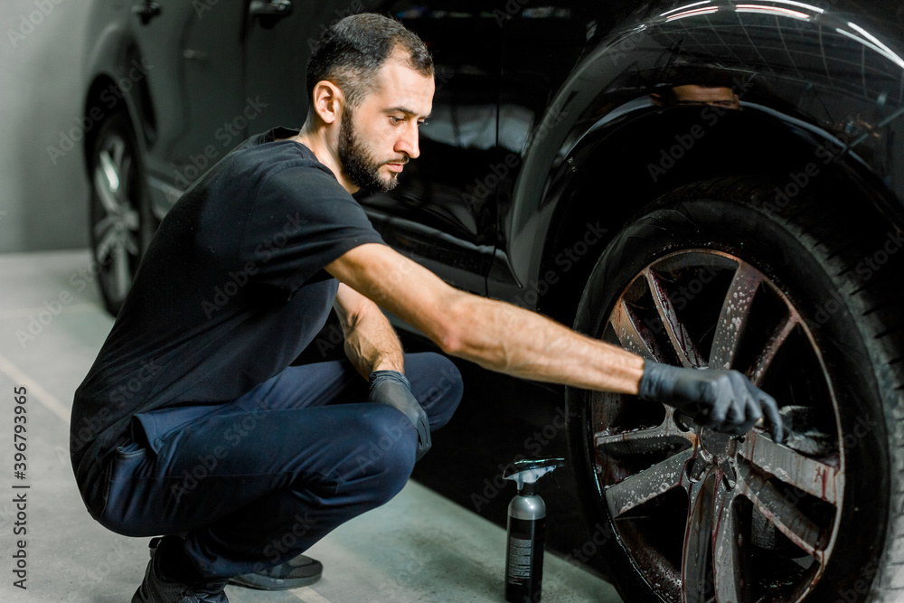Close up of handsome young male employee worker cleaning the wheel rim of modern black crossover car by scrubbing brush. Manual wash at car wash maintance service. Close up of handsome young male employee worker cleaning the wheel rim of modern black crossover car by scrubbing brush. Manual wash at car wash maintance service.