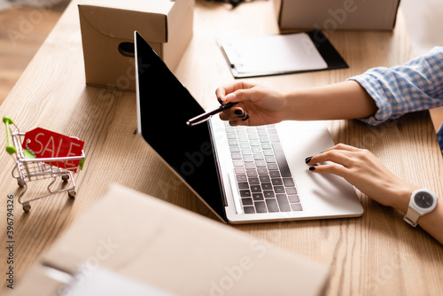Cropped view of volunteer pointing at laptop with blank screen near toy shopping cart with sale lettering and boxes on table