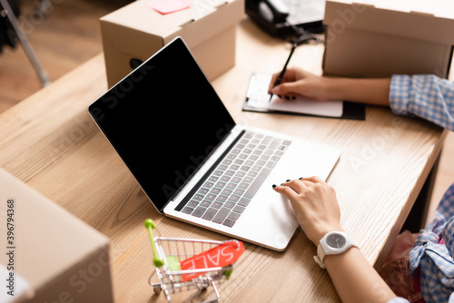 Cropped view of woman using laptop and writing on clipboard near toy shopping cart with sale lettering and boxes in charity center