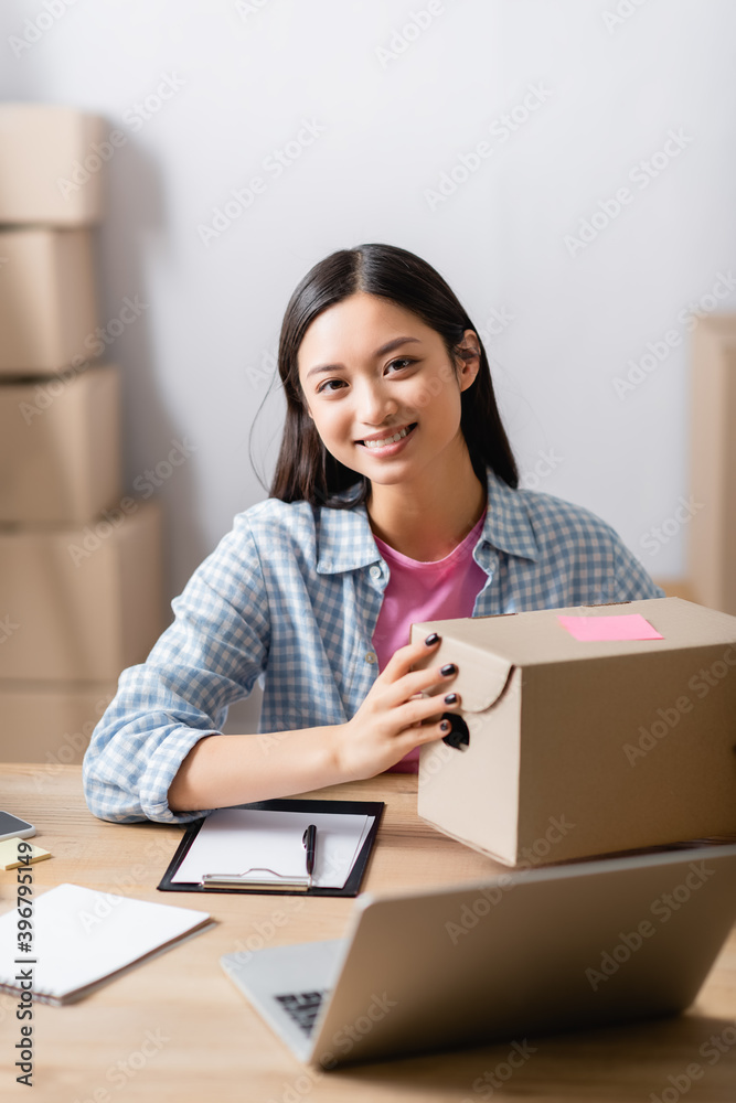 Asian volunteer looking at camera while holding box near clipboard and laptop on blurred foreground Asian volunteer looking at camera while holding box near clipboard and laptop on blurred foreground