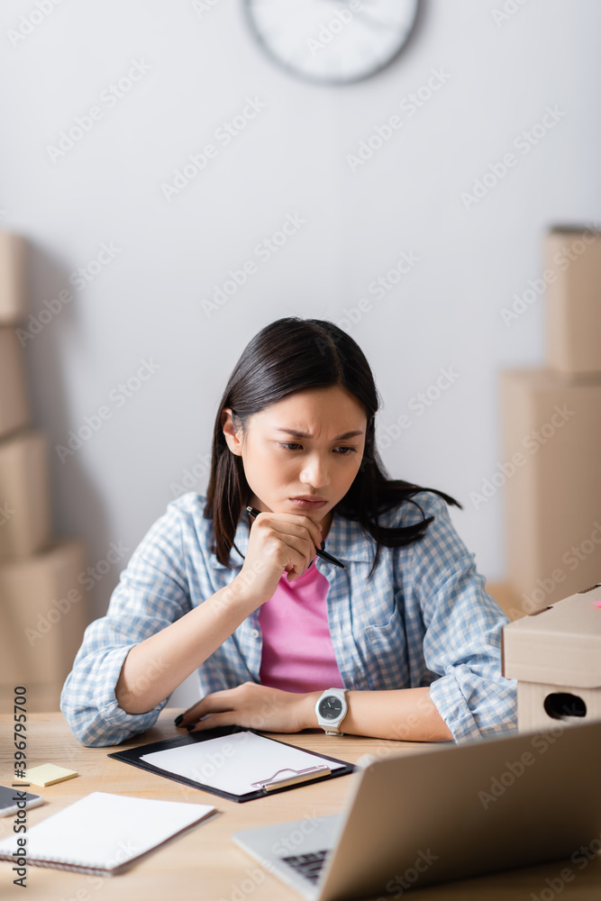 Pensive asian volunteer holding pen and looking at laptop near box and clipboard on blurred foreground Pensive asian volunteer holding pen and looking at laptop near box and clipboard on blurred foreground
