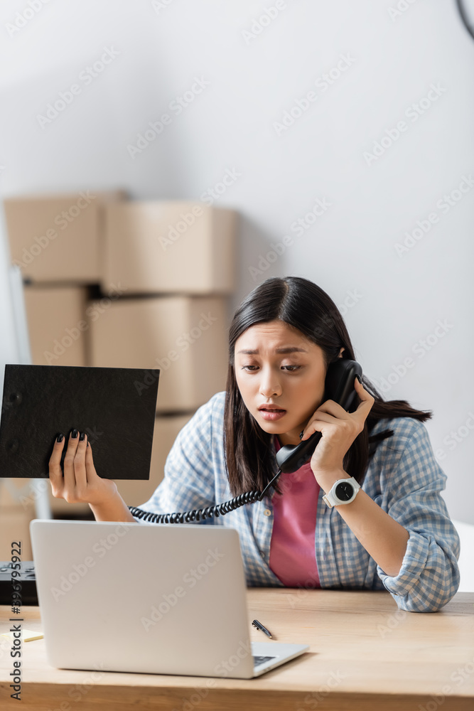 Worried asian volunteer talking on telephone while holding clipboard near laptop on blurred foreground Worried asian volunteer talking on telephone while holding clipboard near laptop on blurred foreground
