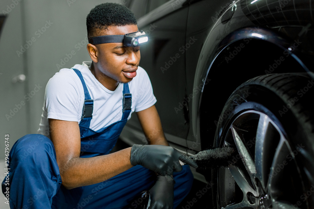 Car washing and detailing workshop. African man worker in protective overalls and rubber gloves, washing car alloy wheel on a car wash, using special brush. Close up portrait, selective focus