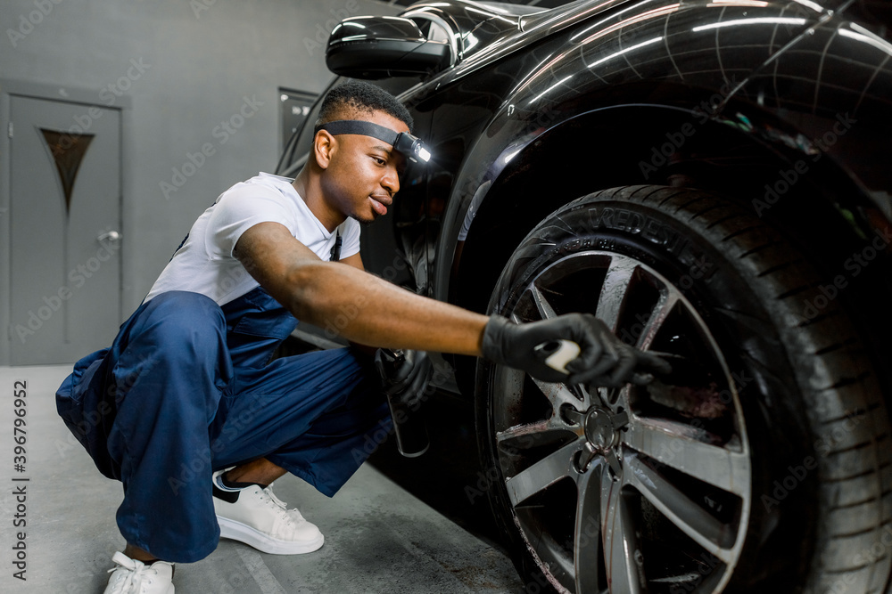 Washing a car by hand, car detailing. Close up image of professional African male worker, cleaning the car wheels with brush. Car rims wash, manual wheel cleaning concept. Washing a car by hand, car detailing. Close up image of professional African male worker, cleaning the car wheels with brush. Car rims wash, manual wheel cleaning concept.