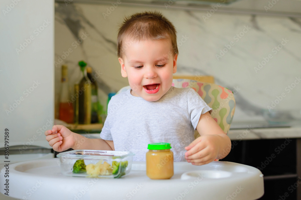 Happy 2-year-old boy kid eats cauliflower, broccoli and mashed potatoes in a jar in his kitchen at home. Baby food, solid food concept. Happy 2-year-old boy kid eats cauliflower, broccoli and mashed potatoes in a jar in his kitchen at home. Baby food, solid food concept.