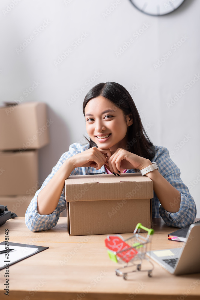 Happy asian volunteer with carton box looking at camera while sitting at desk in charity center on blurred foreground Happy asian volunteer with carton box looking at camera while sitting at desk in charity center on blurred foreground