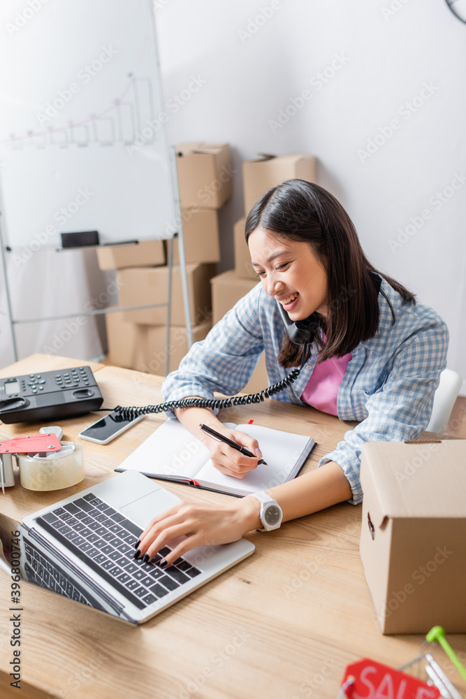 Smiling asian volunteer with pen, talking on telephone while typing on laptop at desk with carton box in charity center Smiling asian volunteer with pen, talking on telephone while typing on laptop at desk with carton box in charity center