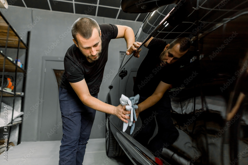 Handsome young Caucasian man, professional worker of car detailing service, polishing black car with microfiber cloth. Car detailing, polishing, finishing concept