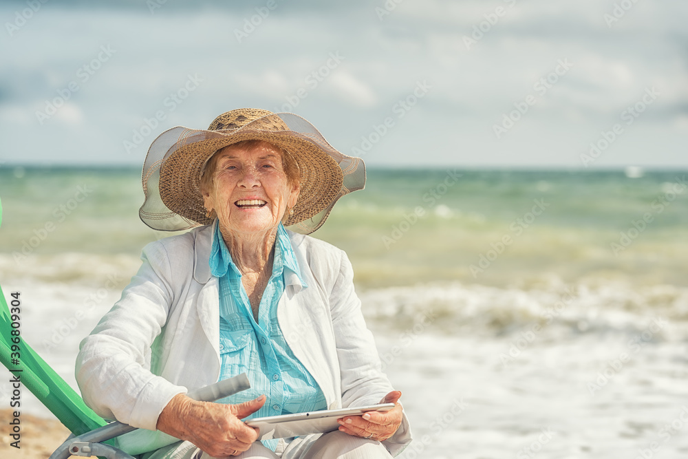 Fototapeta premium Happy laughing old woman 84 years old sitting on the seashore with a tablet computer in her hands.
