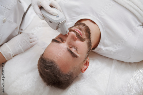 Close up of a bearded man enjoying getting laser facial treatment by professional cosmetologist