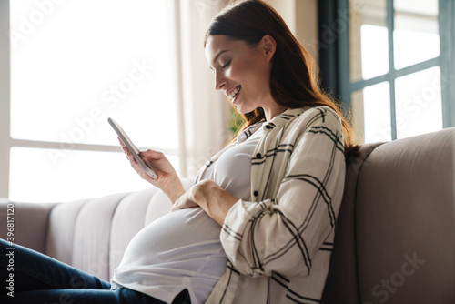 Happy charming pregnant woman using mobile phone while sitting on sofa photo