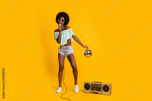 Full length body size photo of black skinned girl performing song with disco ball at concert with boombox isolated on vibrant yellow color background