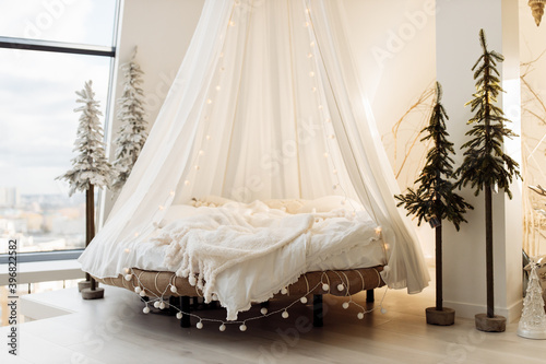 Photo of canopy bed with soft blanket and light garland, near little Christmas tree. Cozy decorated bedroom. Prepare to winter holidays, xmas background, interior design concept