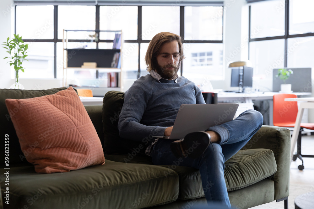 Obraz premium Caucasian businessman using laptop sitting on sofa in creative office