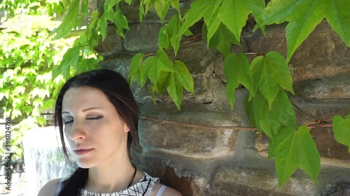 Young brunette woman sitting and resting in park near fountain under the wall with green leafs