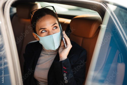 Pleased businesswoman wearing face mask talking on mobile phone in car
