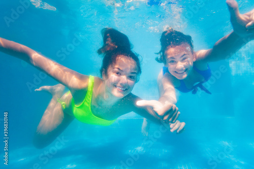 Two happy teenage girls swim playing together underwater in the pool waving hands showing cheerful hand gestures
