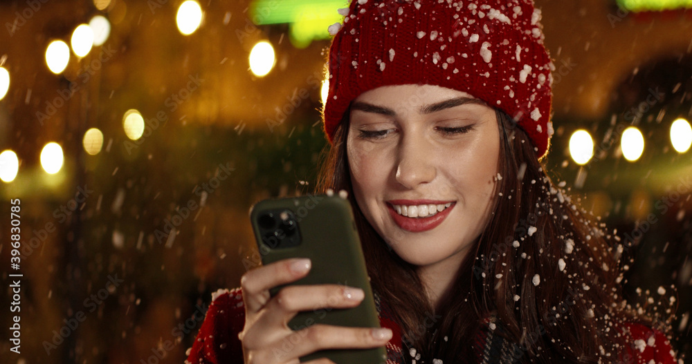 Close up portrait of cheerful Caucasian beautiful female in mask browsing on smartphone while standing on crowded snowy street. Joyful young woman with christmas present texting on cellphone Close up portrait of cheerful Caucasian beautiful female in mask browsing on smartphone while standing on crowded snowy street. Joyful young woman with christmas present texting on cellphone