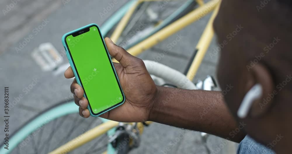 Close up of African American Man in Earphones Holding his Smartphone Looking to Green Screen. He Sitting on Steps in the City with Stylish Colourful Bike at the Background.