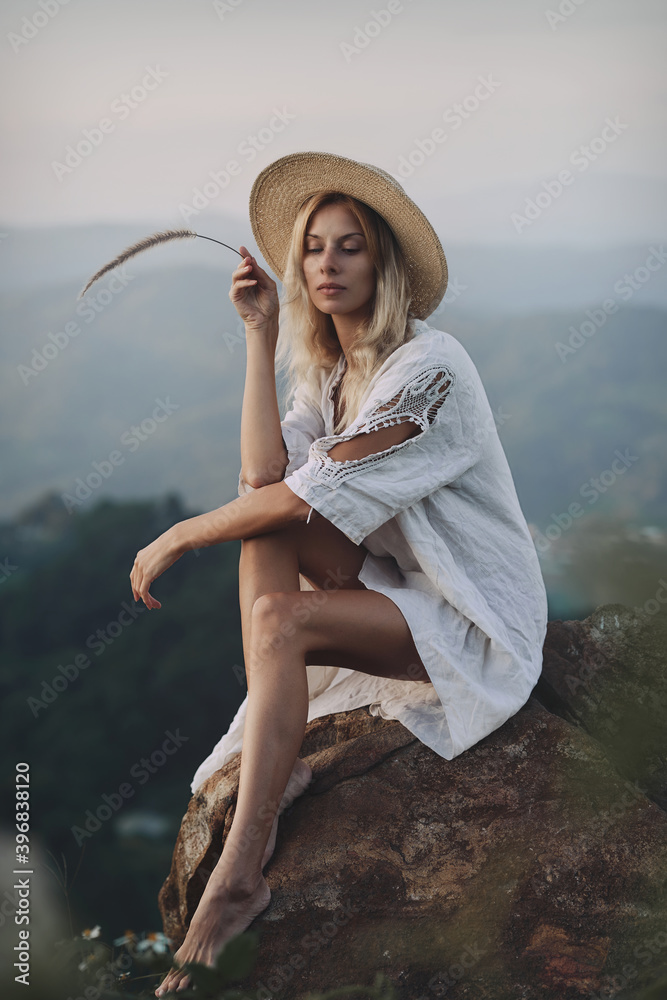 The blonde in a linen dress and straw hat sits on a viewpoint in the mountains. A woman in the Boho style, resting in the countryside, a simple village lifestyle