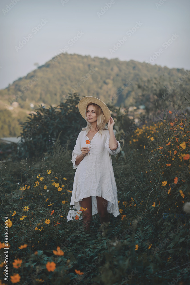 The blonde in a linen dress and straw hat walks among herbs and wildflowers on a meadow in the mountains. A woman in the Boho style, resting in the countryside, a simple village lifestyle
